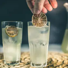 Dłoń dodaje plasterek suszonej limonki do orzeźwiającego kordiału z lodem. Obok stoi drugi kieliszek i dzbanek z napojem.