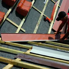 Budowa dachu: pracownik montuje blaszane elementy, obok stosy dachówek czekają na ułożenie. To kluczowy etap prac przy koszu dachowym lukarny.