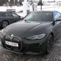 Zielone BMW i4 na ładowarce, obok ciemny Porsche Taycan. Zimowy krajobraz i ślady opon na mokrej nawierzchni.