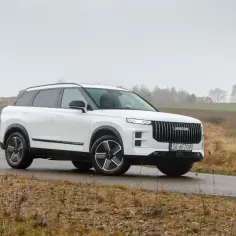 Biały Jaecoo 7 Super Hybrid na mokrej drodze, w tle wiejski krajobraz.