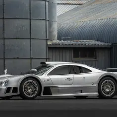 Srebrny Mercedes CLK GTR, ikona wyścigów, zaparkowany przed industrialnymi budynkami.