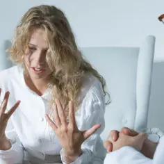 Kobieta w białej koszuli, wyrażająca emocje, rozmawia z lekarzem. Może to być sesja terapeutyczna, gdzie omawiane są leki, np. klozapina.