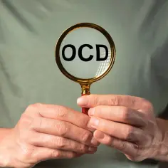 Dłonie trzymają lupę, przez którą widać napis OCD. Może to symbolizować potrzebę zrozumienia zaburzeń obsesyjno-kompulsywnych, w leczeniu których stosuje się klomipraminę.