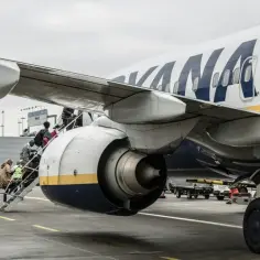 Pasażerowie z bagażem wchodzą po schodkach do samolotu Ryanair. Słońce przebija się przez chmury, zapowiadając podróż.