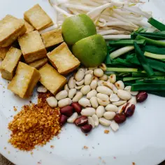 Składniki do sosu pad thai: smażone kostki tofu, orzeszki ziemne, szczypiorek, kiełki fasoli mung, limonki i płatki chili.