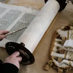 Osoba wskazuje na tekst na zwoju Tory, który jest rozłożony na stole w synagodze. Obok leży ozdobny pokrowiec.