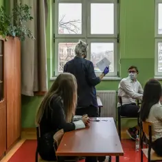 Nauczyciel dyplomowany zarobki - lekcja w klasie, uczniowie w maskach siedzą przy ławkach, nauczyciel trzyma kartkę.