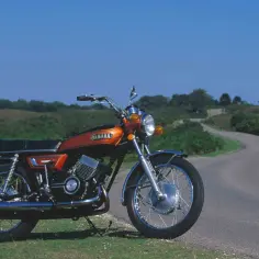 Pomarańczowy motocykl Yamaha RD 350 na tle krętej drogi i zielonych wzgórz. Klasyk gotowy na podróż.