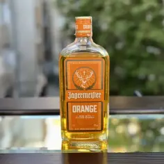 Butelka Jägermeister Orange, z pomarańczowym akcentem, stoi na lustrzanym blacie, w tle zieleń.