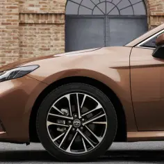 Elegancka, brązowa Toyota Camry 2024 z nowymi światłami i felgami, zaparkowana przed ceglanym budynkiem.