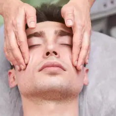 Mężczyzna z zamkniętymi oczami doświadcza relaksującego masażu twarzy podczas kursu Kobido. Dłonie terapeuty delikatnie głaszczą jego skórę.