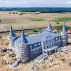 Budowa hotelu 4 wieże w kształcie zamku, otoczonego polami i lasem. Widać plac budowy z maszynami i materiałami.
