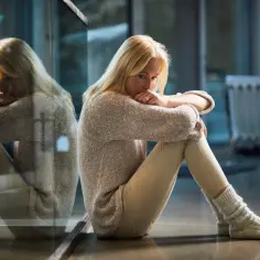 Blondynka siedzi przy oknie, pogrążona w myślach. Jej odbicie w szybie podkreśla uczucie samotności.