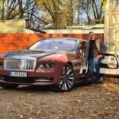 Elegancki mężczyzna obok luksusowego Rolls-Royce'a elektrycznego w kolorze bordowym. Jesienne liście wokół.