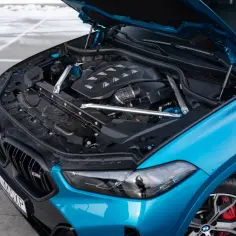 Błękitne x6 BMW z otwartą maską, odsłaniającą potężny silnik M. Widoczne detale podkreślają sportowy charakter auta.