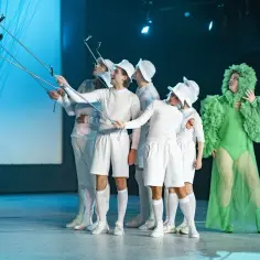 Aktorzy w białych strojach robią sobie selfie na scenie Teatru Capitol Poznań. Obok nich stoi postać w zielonej, falbaniastej kreacji.