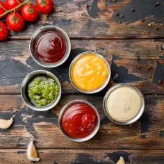 Pięć sosów do grilla: ketchup, musztarda, sos czosnkowy, sos barbecue i zielony sos. Obok pomidory i czosnek.