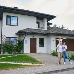 Rodzina przed nowoczesnym domem. Ojciec niesie córkę na barana, matka obok. W tle biały dom z brązowymi drzwiami i garażem. To ich wymarzony dom, na który przygotowali szczegółowy kosztorys.
