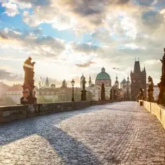 Kamienny most z rzeźbami i latarniami prowadzi do zabytkowego miasta w Czechach. Słońce wschodzi nad kopułami i wieżami.