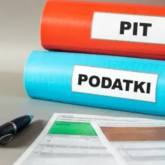 Dwa segregatory: czerwony z napisem "PIT" i niebieski z napisem "PODATKI". Obok leży długopis i formularze podatkowe.