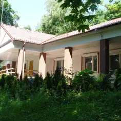 Dom z werandą i ogrodem, otoczony zielenią. W tle drzewa. Krynica Morska rynek.