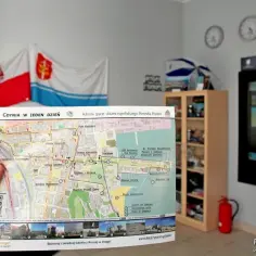 Ręka trzyma mapę "Gdynia w jeden dzień", pokazującą trasę spaceru po Gdyni. W tle flagi i zegary.