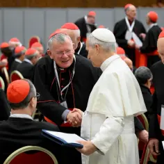 Papież Franciszek podczas konsystorza ściska dłoń kardynałowi. W tle inni kardynałowie w czerwonych biretach.