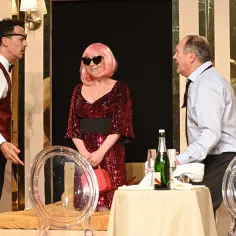 Trzech aktorów na scenie Teatru Capitol Kraków. Kobieta w błyszczącej sukni i peruce, mężczyźni w eleganckich strojach.