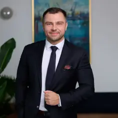 Mężczyzna w garniturze, stojący obok rośliny, z obrazem w tle.