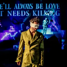 Mężczyzna w oliwkowej marynarce z zawiązanymi oczami na scenie. W tle napisy "THERE'LL ALWAYS BE LOVE THAT NEEDS KILLING". Spektakl w Warszawie.