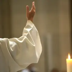 Kapłan w białej szacie podnosi rękę. Czy opuszczając jedną mszę można iść do komunii? Płonąca świeca obok.