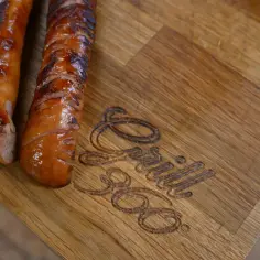 Dwie soczyste kiełbaski z grilla leżą na drewnianej desce z wygrawerowanym logo "Grill 360".