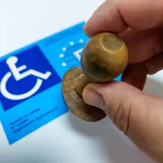 Dłoń trzyma pieczątkę nad niebieską kartą parkingową z symbolem wózka inwalidzkiego.