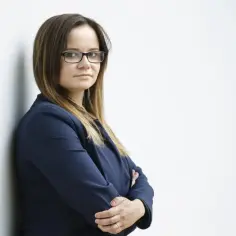 Kobieta w okularach i granatowej marynarce stoi oparta o ścianę, ze skrzyżowanymi rękami.