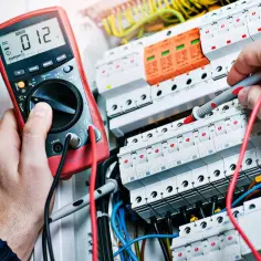 Elektryk w pomarańczowym kasku sprawdza instalację elektryczną multimetrem. To pierwszy krok, jak zostać elektrykiem.