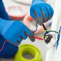 Elektryk w niebieskich rękawicach sprawdza przewody przy gniazdku. Pokazuje, jak podłączyć gniazdko bez uziemienia, używając miernika.