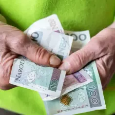 Dłonie trzymają polskie banknoty: 10, 20 i 100 zł. Może to symbolizować wsparcie finansowe, np. renta socjalna dla bezrobotnego.