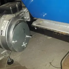 Metalowa klapka regulacyjna na dmuchawie z niebieskim zbiornikiem w tle.