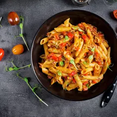 Makaron penne z mięsem mielonym i papryką w czarnej misce, ozdobiony kiełkami i pomidorkami.