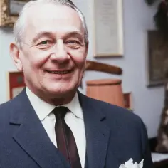 Uśmiechnięty Mieczysław Fogg w garniturze, jakby żył podwójnym życiem, z nagrodami na ścianie.