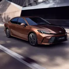 Brązowa Toyota Camry 2.5 Hybrid sunie po drodze, otoczona nowoczesną architekturą i cieniem drzew.