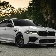 Białe BMW M5 Competition z czarnymi felgami i detalami, zaparkowane na tle drzew i palm.