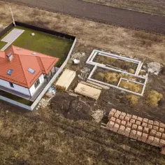 Działka zagrodowa w budowie: gotowy dom z czerwonym dachem, fundamenty, drewno i palety cegieł.