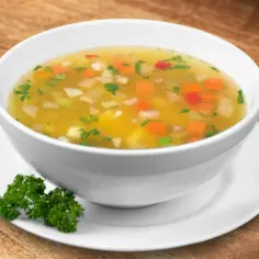 Aromaticzna jarzynowa zupa z kawałkami marchewki, ziemniaków i zielonej pietruszki w białej misce na drewnianym tle.
