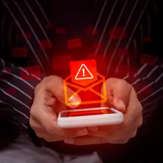 Dłonie trzymają smartfon, z którego wychodzi ostrzeżenie o nieznanym nadawcy. Wokół unoszą się czerwone koperty.