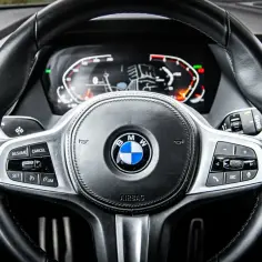 Kierownica BMW z cyfrowym kokpitem i przyciskami tempomatu. HBA (asystent świateł drogowych) włączony, widoczny na ekranie.