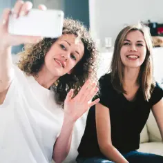 Dwie przyjaciółki robią sobie selfie. Jedna z nich, z kręconymi włosami, macha do aparatu. Zastanawiamy się, czyje są dzisiaj imieniny.