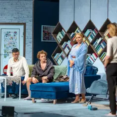 Na scenie inteligenci spektakl. Mężczyzna w białej koszuli, młody chłopak w bluzie i kobieta w niebieskiej sukience siedzą i stoją, otoczeni książkami.