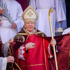 Biskup w czerwonej szacie i mitrze, trzymający pastorał, otoczony przez ministrantów.
