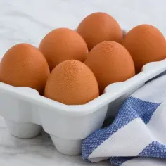 Sześć brązowych jajek w białym wytłaczanym pojemniku, obok niebiesko-biała ściereczka.